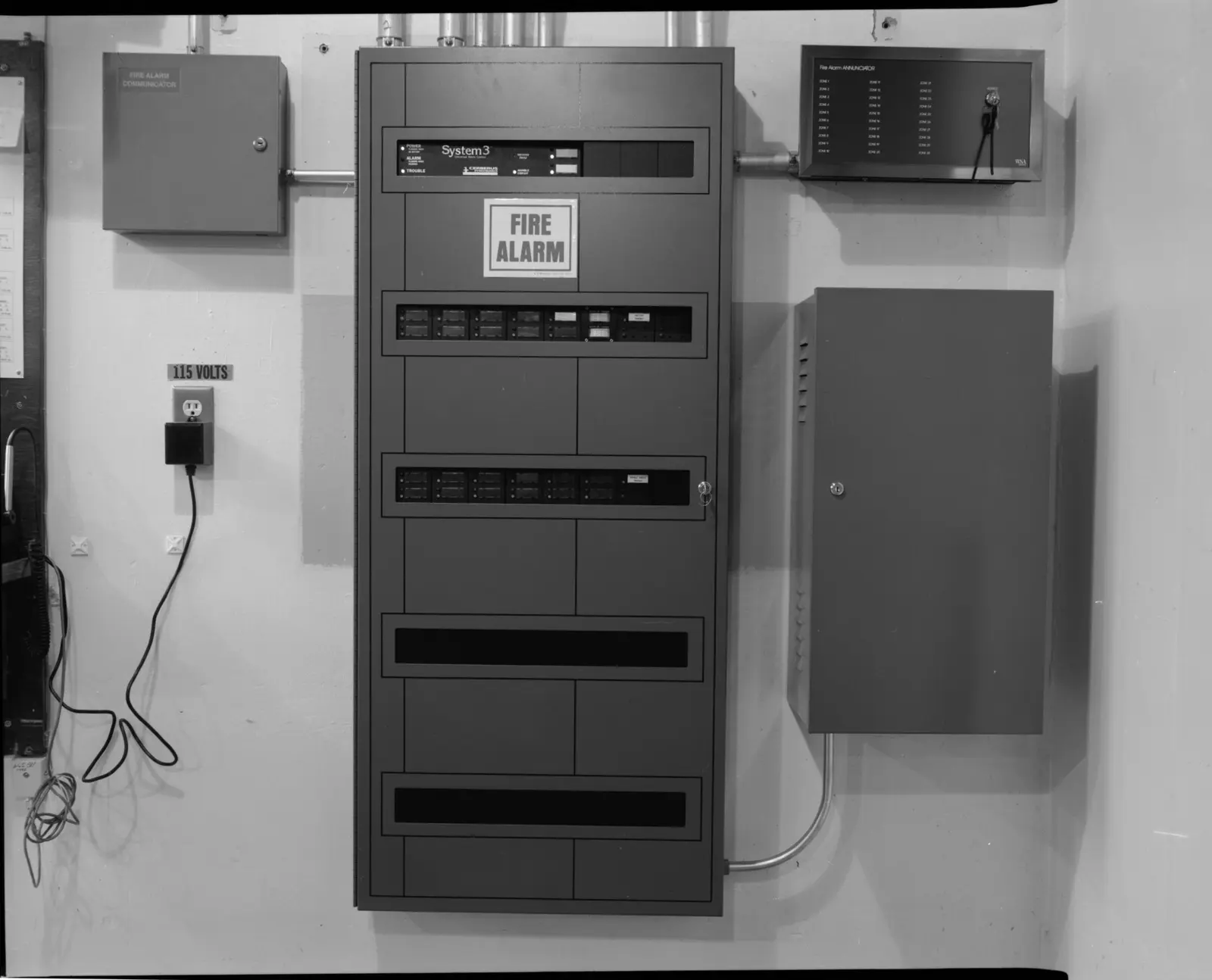 An older commercial fire alarm control panel in a utility room, with a separate fire alarm communicator box on the left and a zone annunciator on the right
