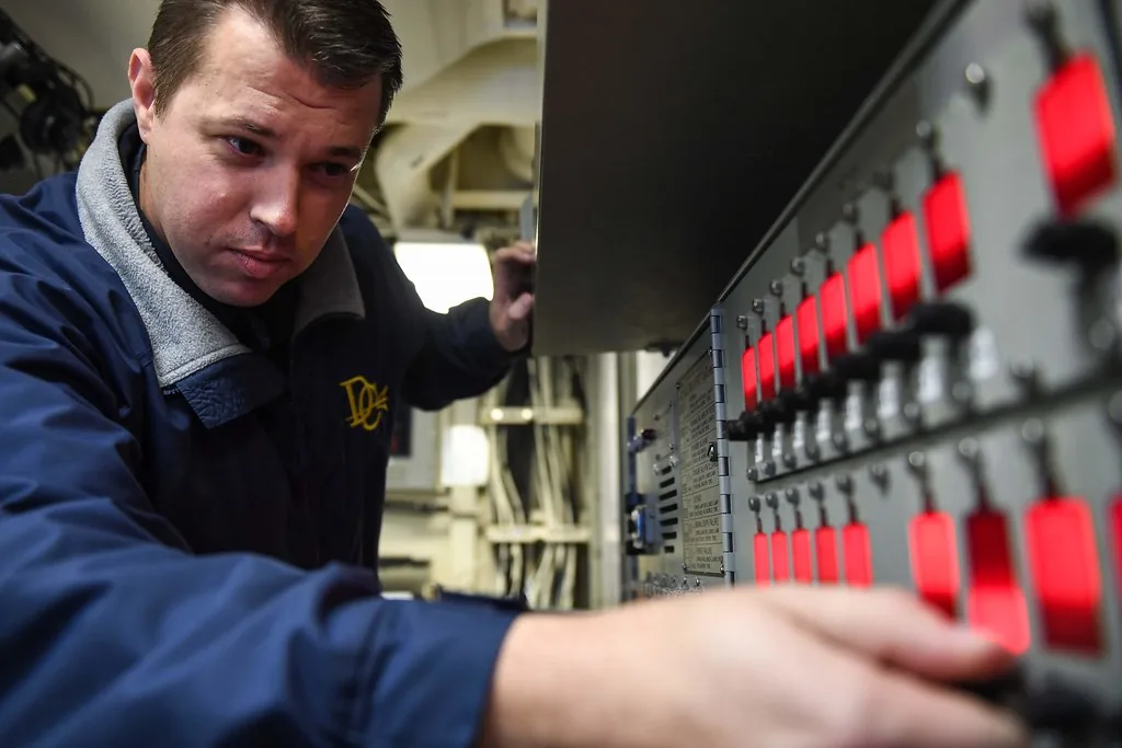 A technician in a blue jacket tests an alarm panel, reaching toward a row of red indicator lights