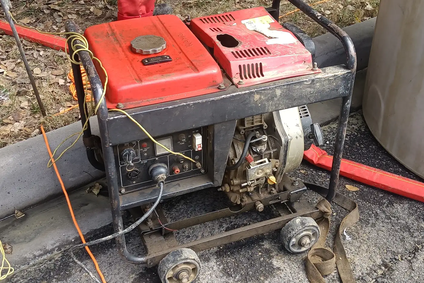 A red portable engine generator with a cord plugged into its control panel, standing on wheels on pavement outdoors