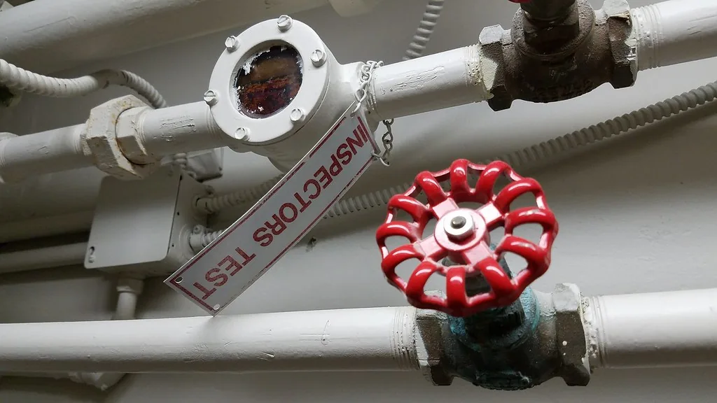Red sprinkler valve wheel on white pipes with a tag reading INSPECTORS TEST